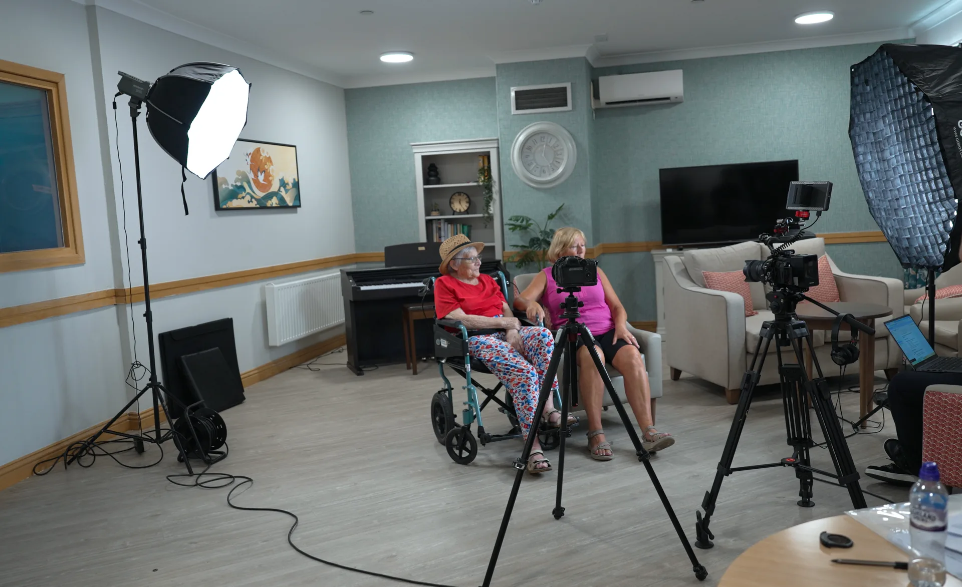 A small crew filming quietly in the lounge while a carer sits with a resident.
