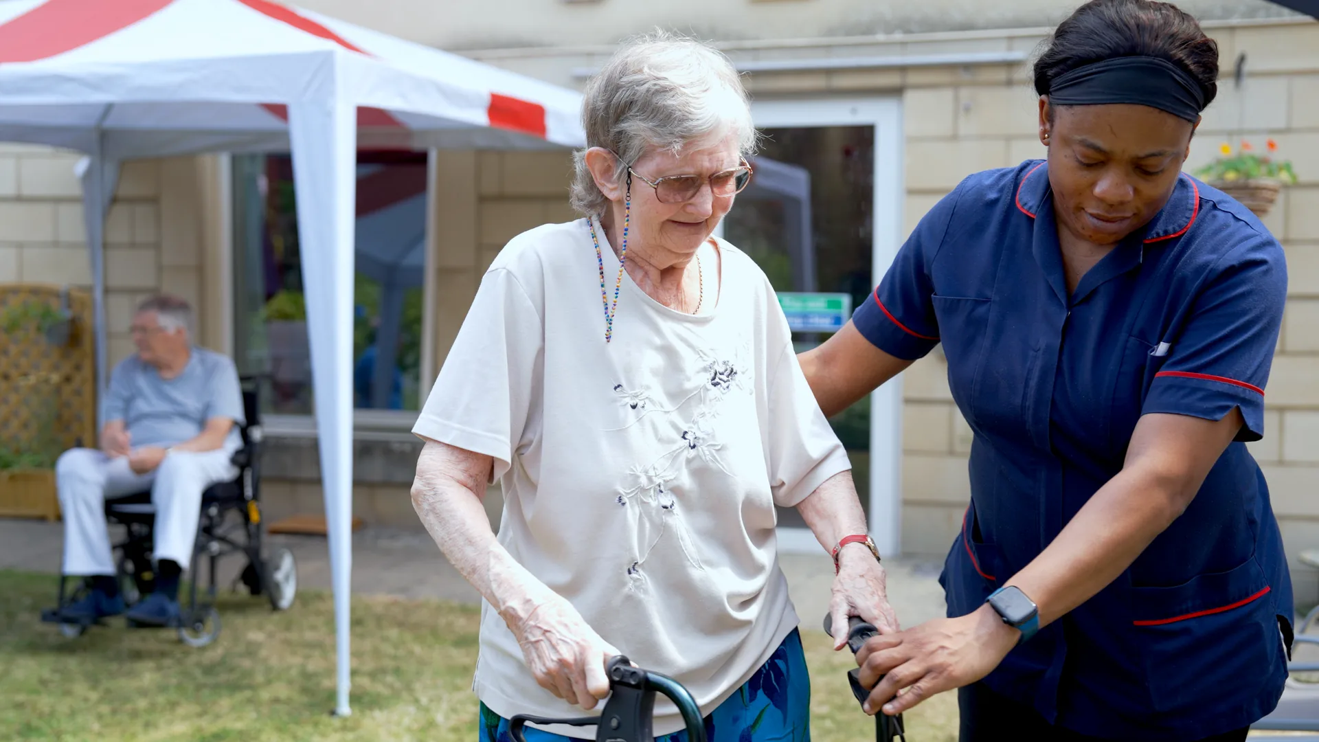 A carer helping a resident walk with her frame in the garden.